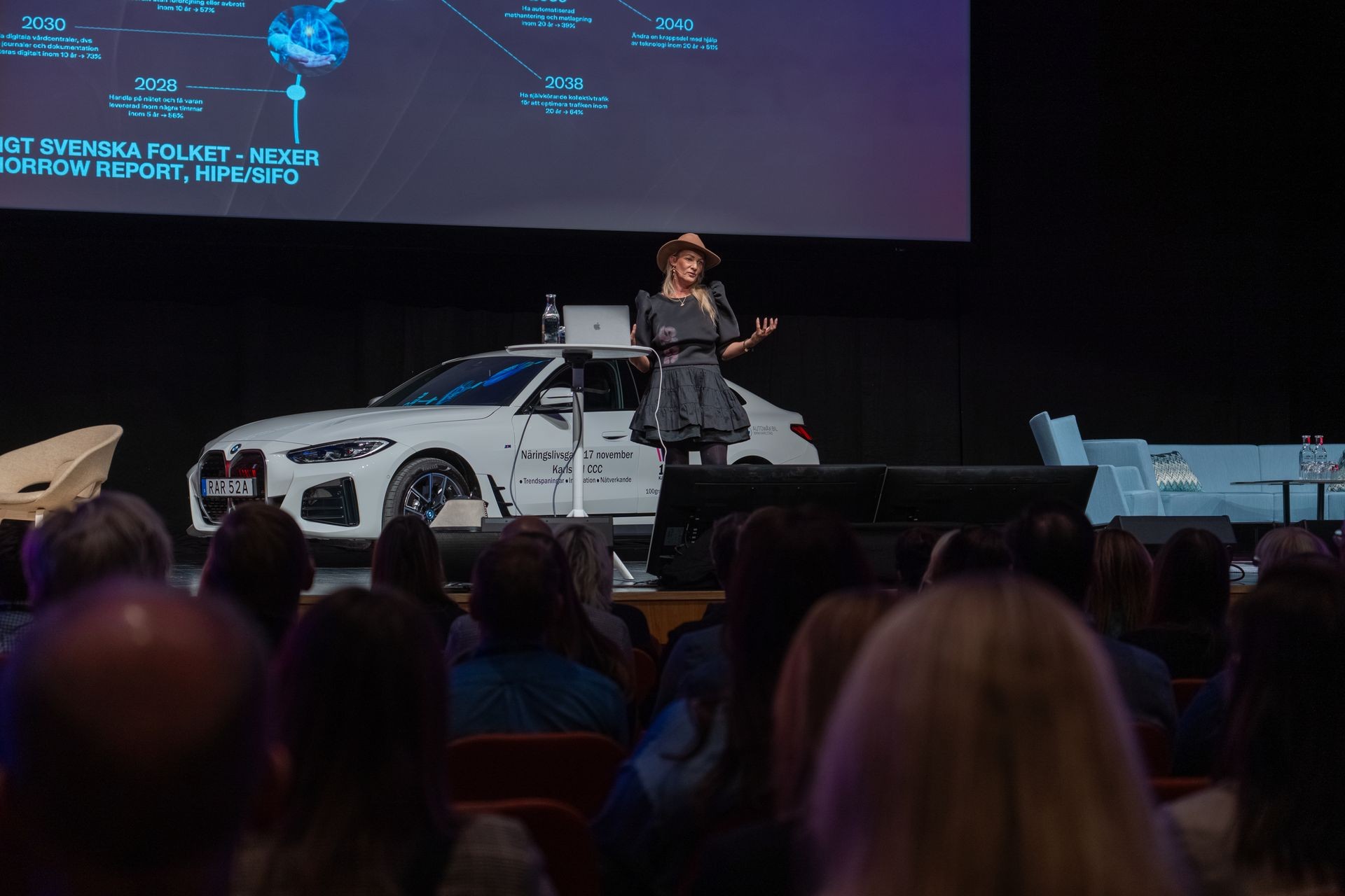 Speaker presenting on stage with a white car in the background and a large screen displaying informational graphics.