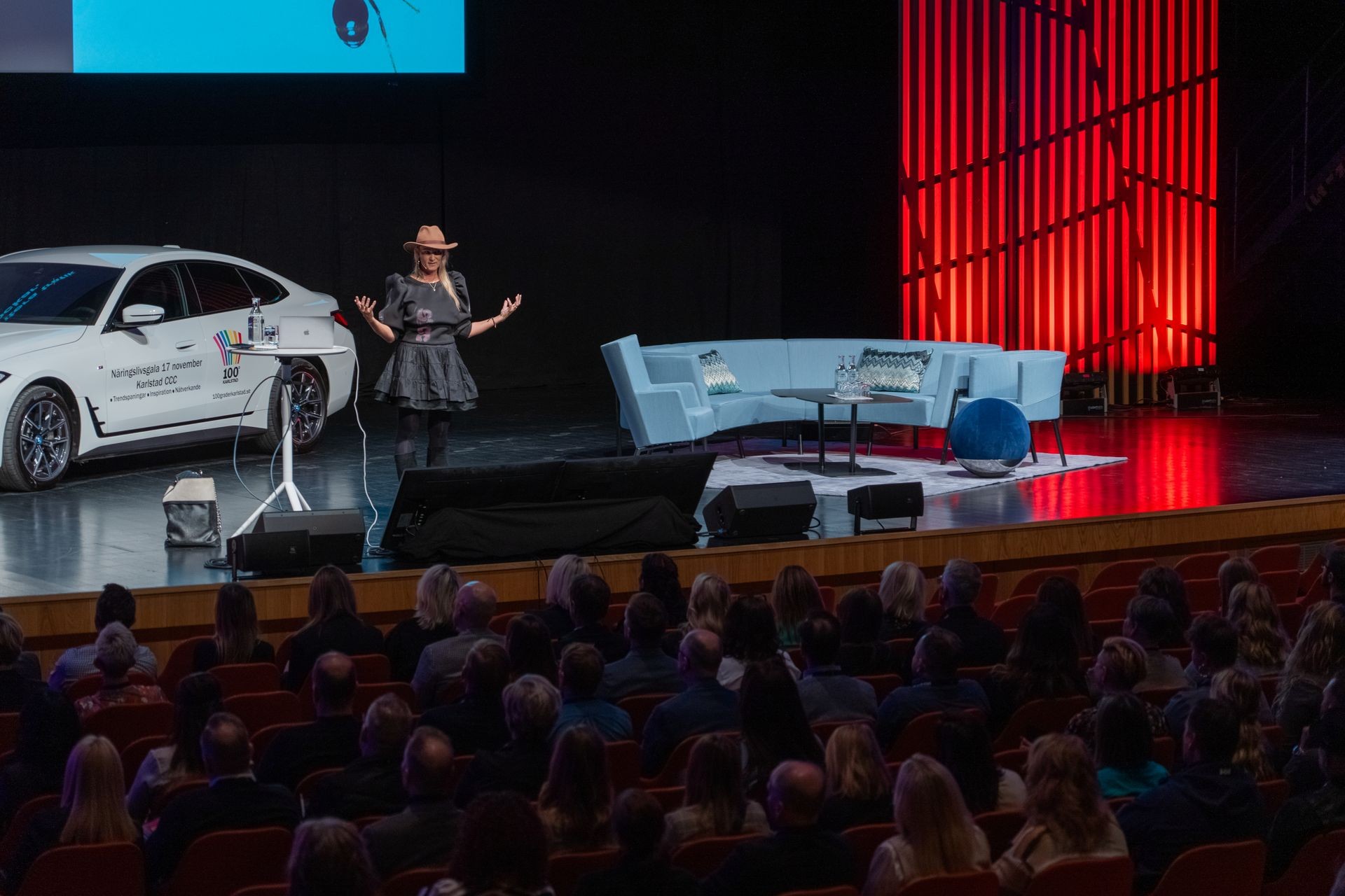 Speaker on stage with a white car, delivering a talk in front of a seated audience.